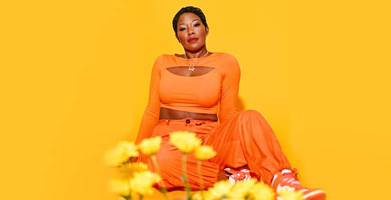 Laci Jordan wears an orange outfit, the background is yellow. A few yellow flowers are visible in the foreground.